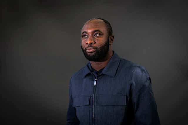 A bearded man in a denim shirt poses for a photo against a dark grey backdrop. He looks into the distance without smiling.