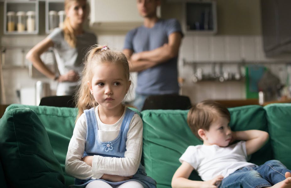 Une jeune fille en colère est assise avec son frère sur le canapé, tandis que leurs parents les observent à l'arrière-plan.