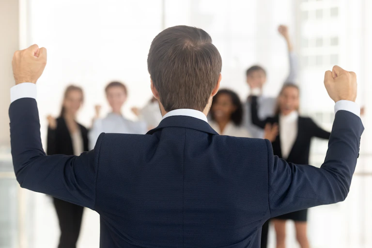 back of man in suit with hands raised, facing a group of people in work clothes with fists in the air.