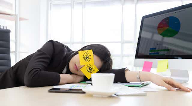 Woman in suit jacket asleep on a desk with eyes drawn on post-its on her face, computer in the background, teacup, phone and papers on the desk.