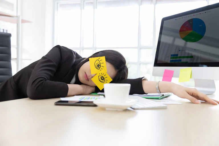 Woman in suit jacket asleep on a desk with eyes drawn on post-its on her face, computer in the background, teacup, phone and papers on the desk.