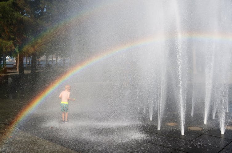 A young boy plays in a fountain, with a rainbow overhead.