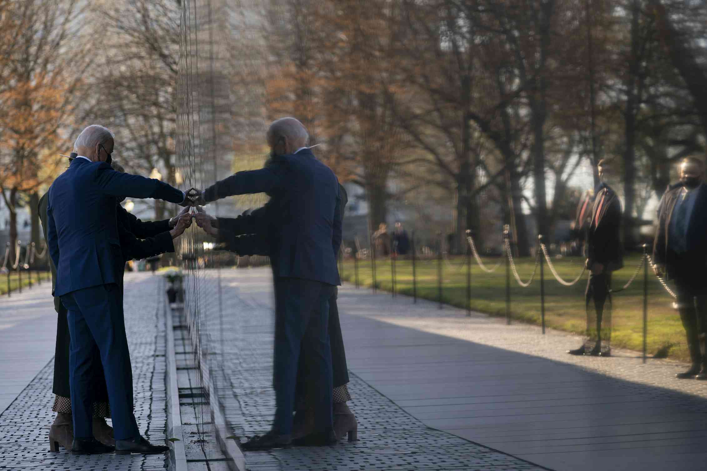 A man and woman touch the etchings of names on a war memorial, their reflections visible in the polished stone.