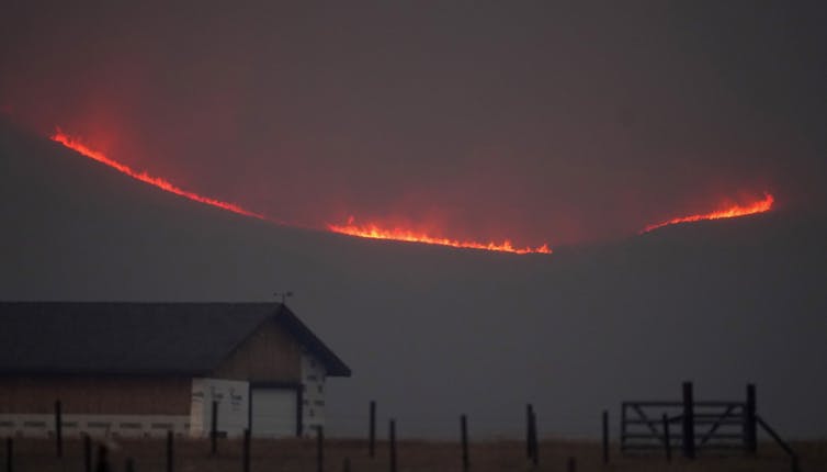 Fire burns in the mountains above a building and ranch fence.