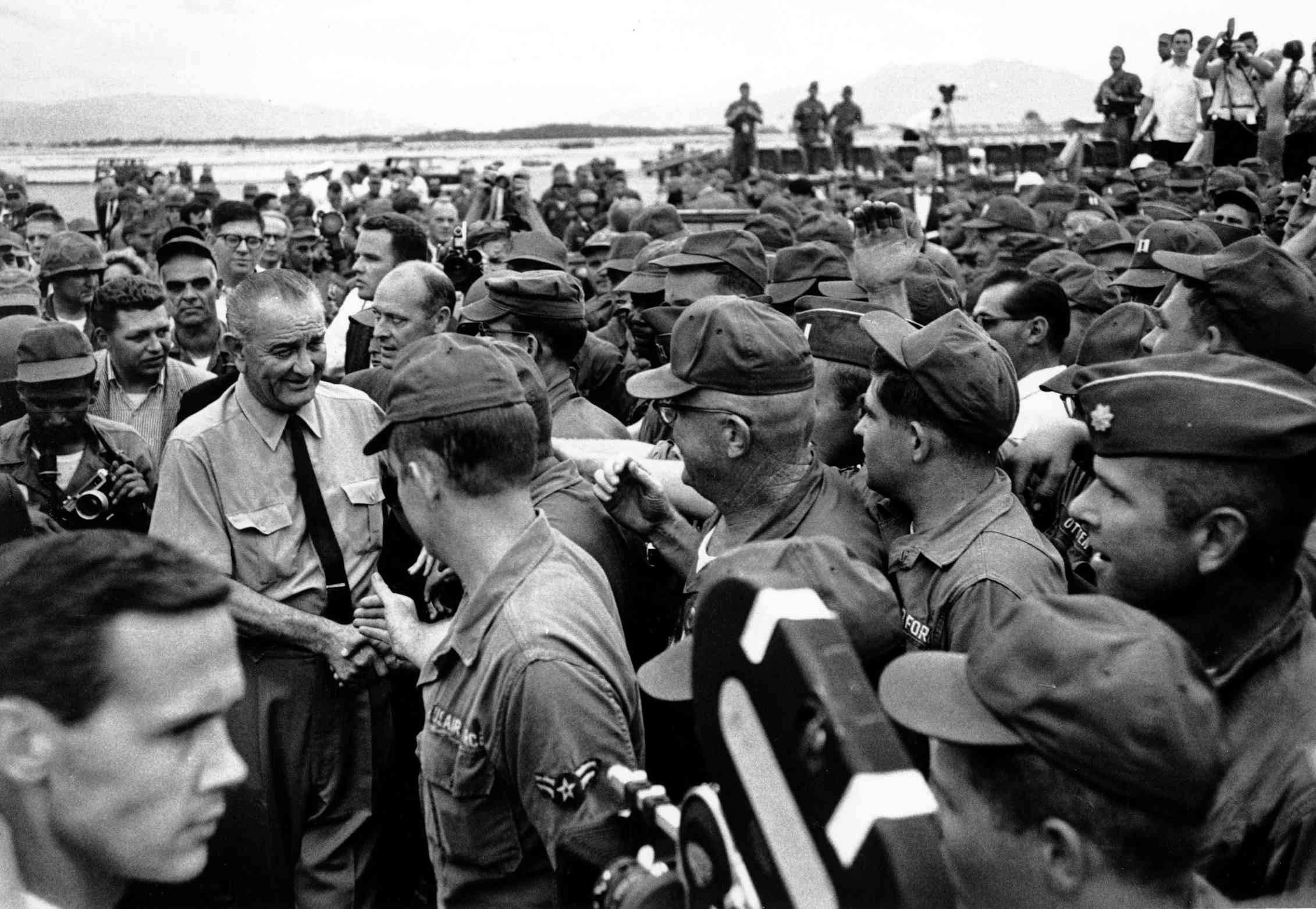 A black and white photo shows an older man in a shirt and tie surrounded by soldiers in uniform.