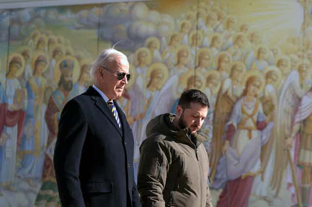 An older man in sunglasses in a dark coat walks next to a younger man in an olive green jacket and pants beside a painted mural with angels.