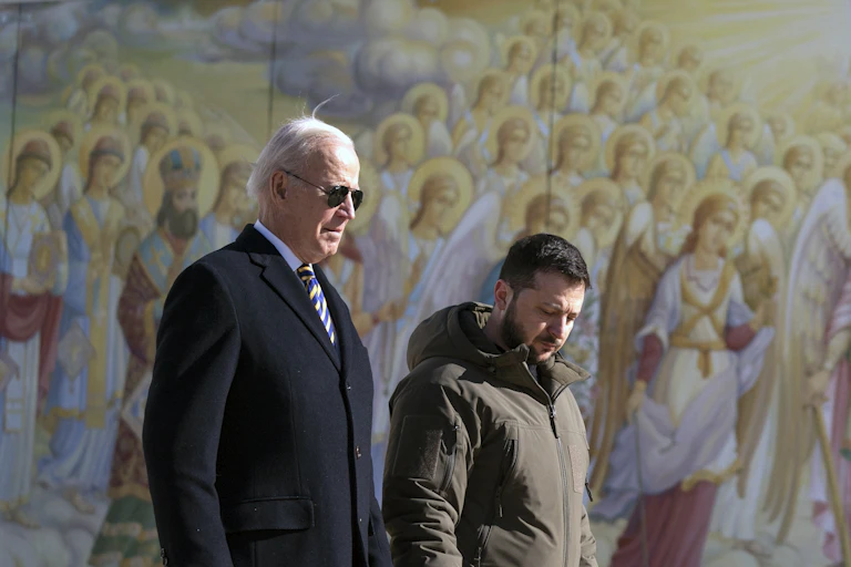An older man in sunglasses in a dark coat walks next to a younger man in an olive green jacket and pants beside a painted mural with angels.