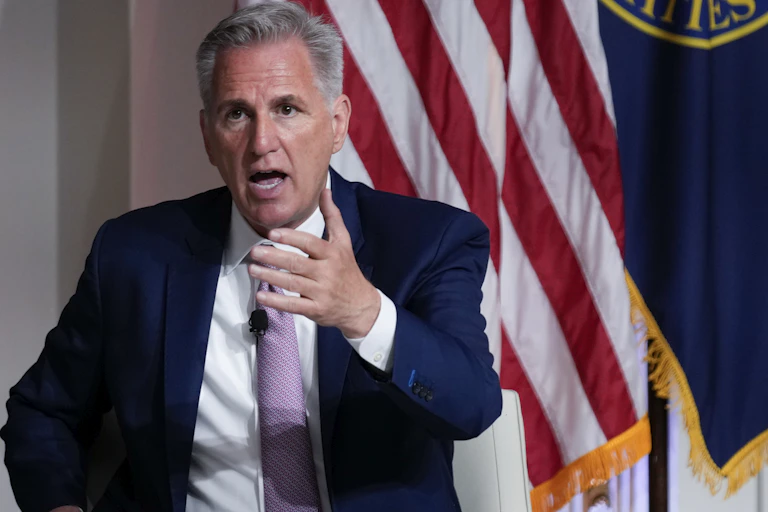 A man in a blue suit and lilac tie gesticulates while standing in front of an American flag.