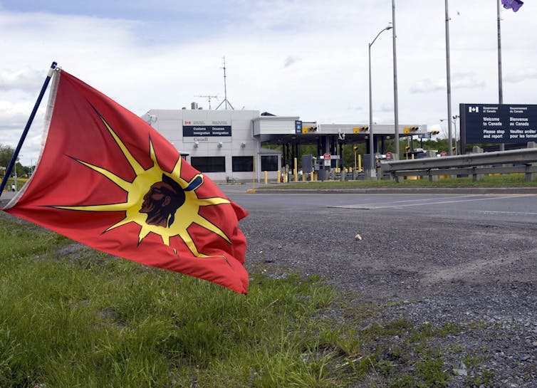 A red and yellow flag with an Indigenous man's profile in the middle flies in front of a Canadian border crossing.