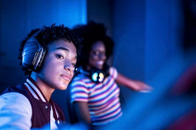 Two young people of color with headphones appear in the blue glare of a computer.