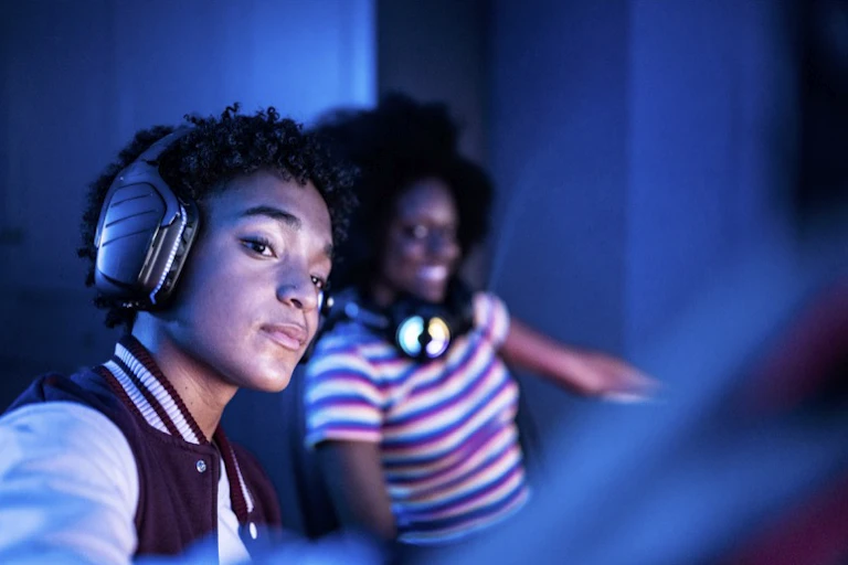 Two young people of color with headphones appear in the blue glare of a computer.