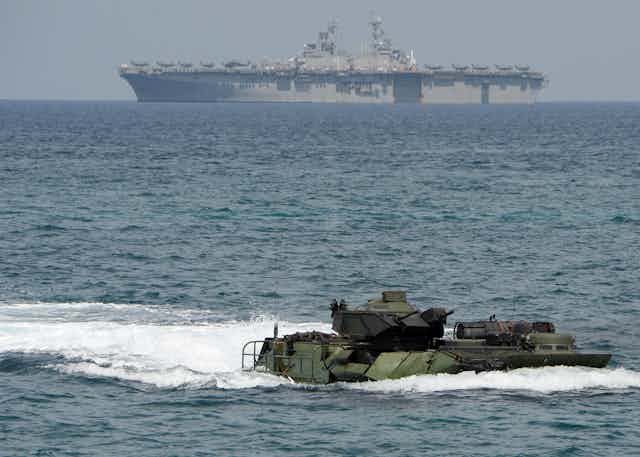 A small vessel in water is seen in the foreground while a huge aircraft carrier is seen in the background.