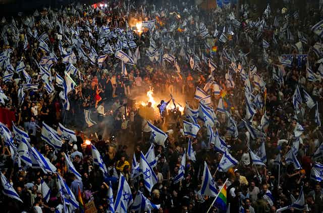 A sea of people carrying white and blue flags surround a bonfire.