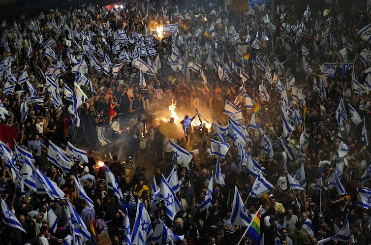 A sea of people carrying white and blue flags surround a bonfire.