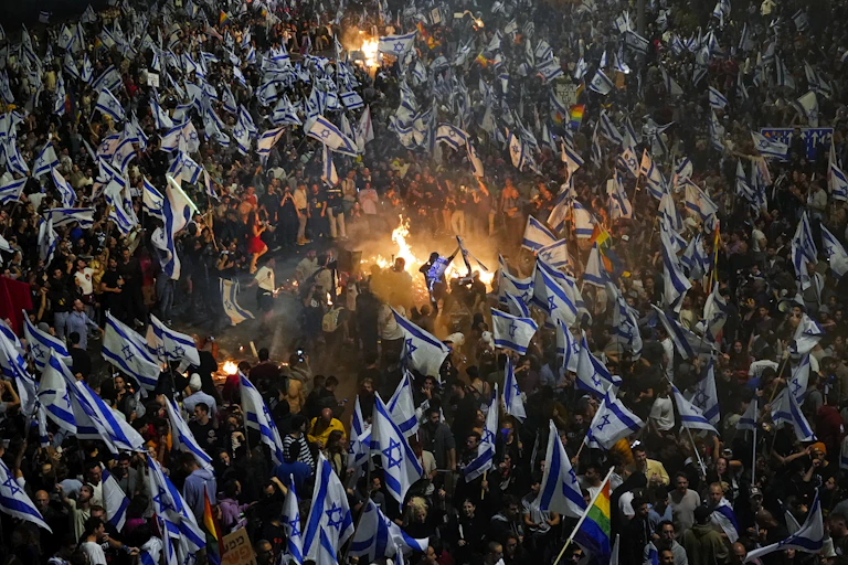 A sea of people carrying white and blue flags surround a bonfire.