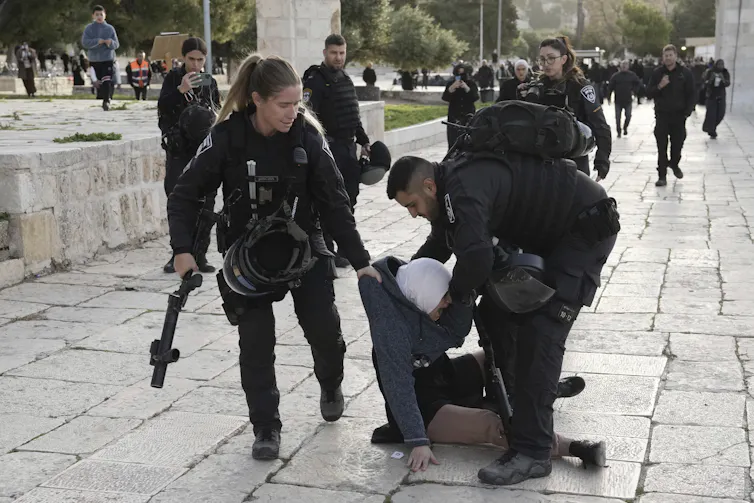 A woman in an hijab sits at the feet of two armed police officers, a man and woman, who appear to be trying to drag her away.