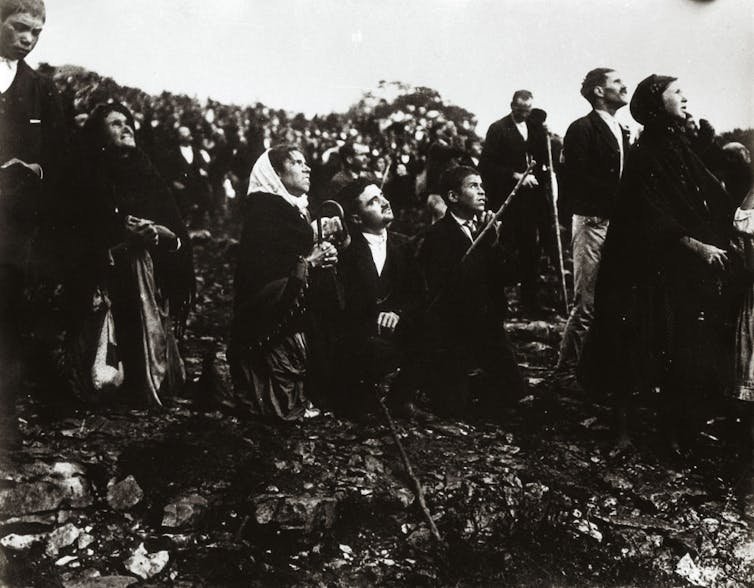 A black and white photograph shows people standing and kneeling in a field, looking up to the sky.