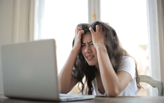 An upset woman with her hands on her head sits in front of a laptop.