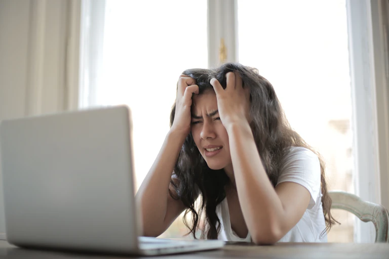 An upset woman with her hands on her head sits in front of a laptop.