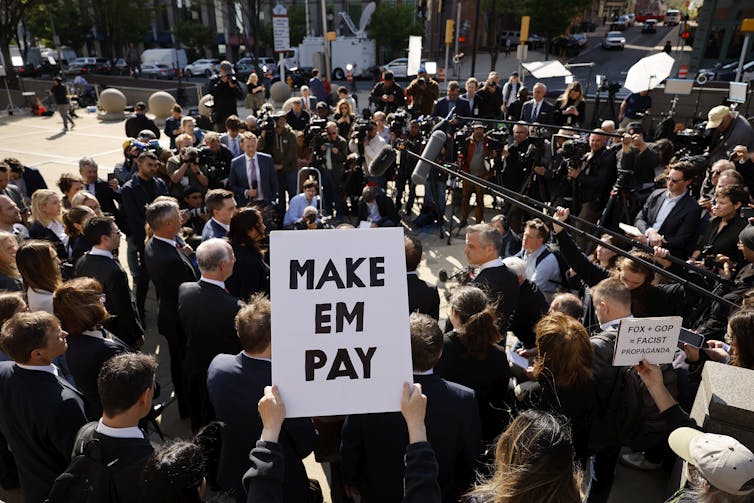 A throng of lawyers speak facing a large group of journalists holding notebooks, microphones and cameras. Someone holds a sign that reads, 