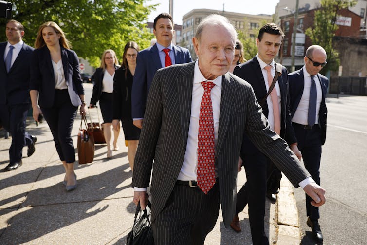 A group of men and women, dressed in suits, some carrying briefcases, all with solemn looks on their faces, cross a city street on a sunny day.