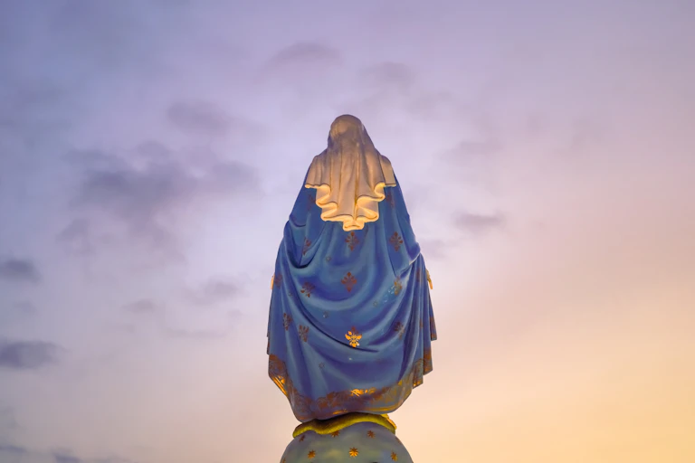A statue of a woman in a blue and gold cloak and white head covering, viewed from the back as the sun rises.