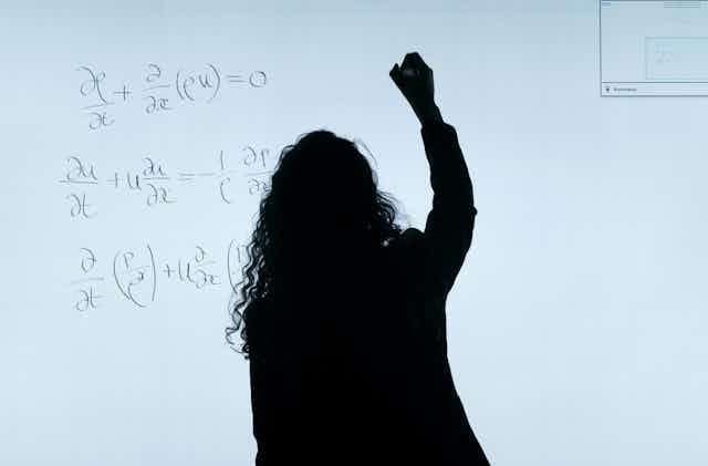 A woman in shadow writes equations on a board.
