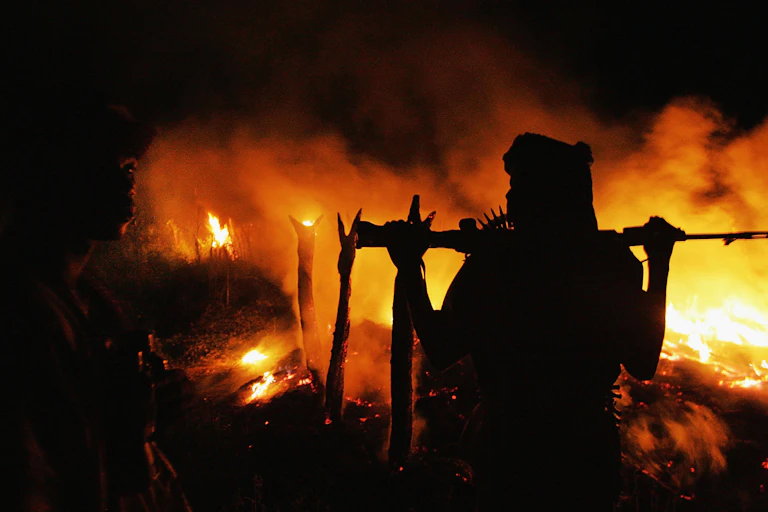A person with a gun stands next to a burning house