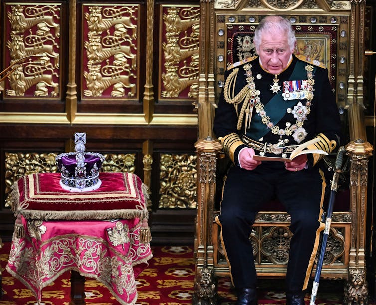 An elderly man slumps on a throne sitting next to a crown embedded with jewels.