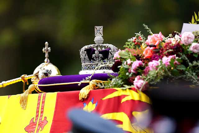 A flag-draped and flower-strewn coffin with a crown and other gem-studded items on top.