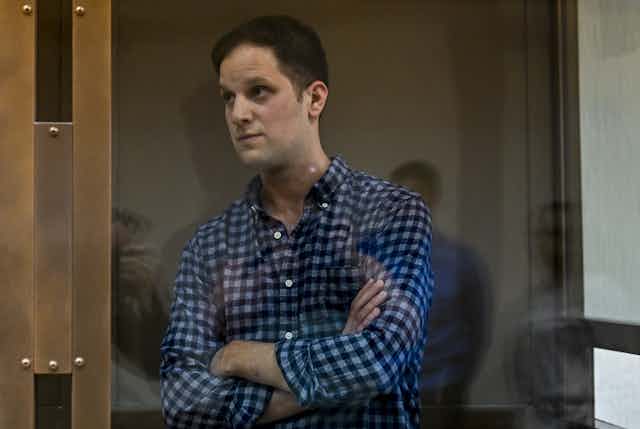 A man in a blue checked shirt stares through some reinforced glass in court.