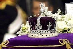 A crown with a large diamond sits on purple velvet atop a coffin.