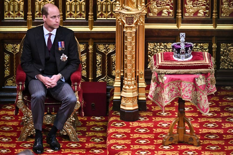 A balding man in a dark suit sits next to an ornate crown encrusted with jewels.