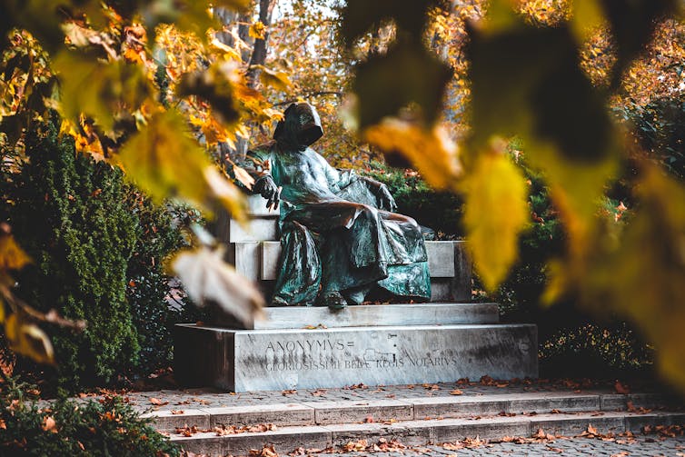 statue of Anonymous in a leafy autumn park. The statue is a hooded figure sat with a pen in their hand.
