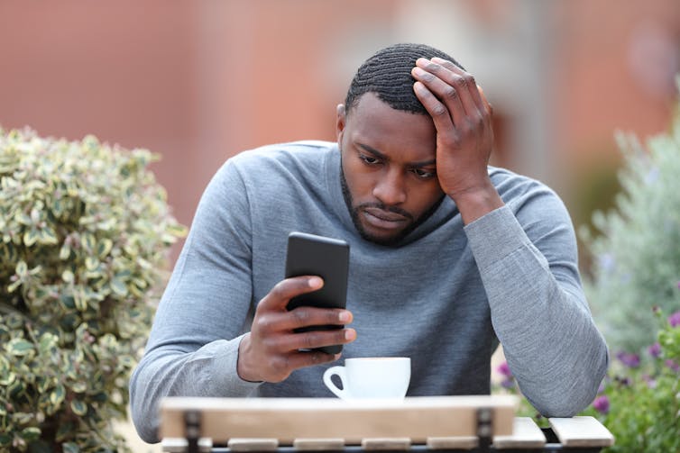 A man sitting at a table looks at his phone with a stressed look on his face.