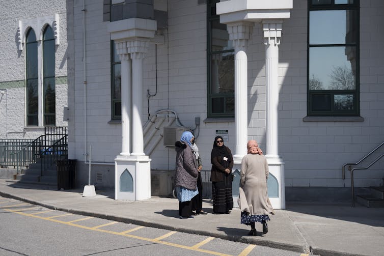 Women wearing hijabs stand on the sidewalk outside a white building.