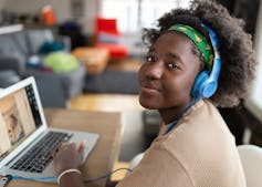 A young African American girl looks toward the camera as she sits at a laptop wearing a pair of blue headphones and a green headband.
