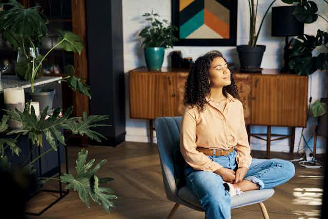 With her eyes closed, a woman dressed in blue jeans and pale orange shirt sits in a chair in her living room engaged in a meditation session.