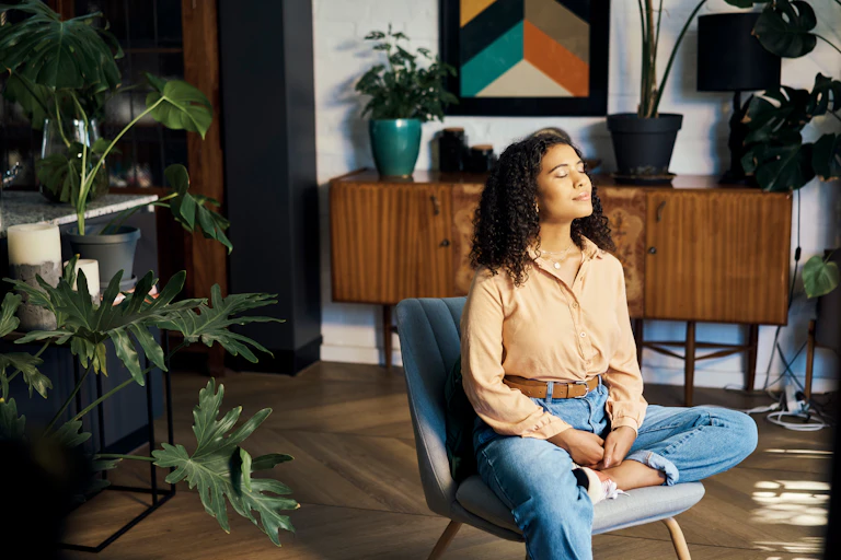 With her eyes closed, a woman dressed in blue jeans and pale orange shirt sits in a chair in her living room engaged in a meditation session.
