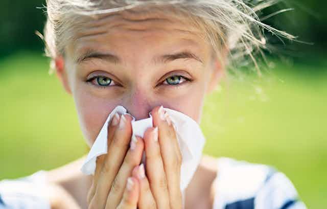 A young woman with allergy-reddened eyes sneezes into a tissue. The background is a lawn.