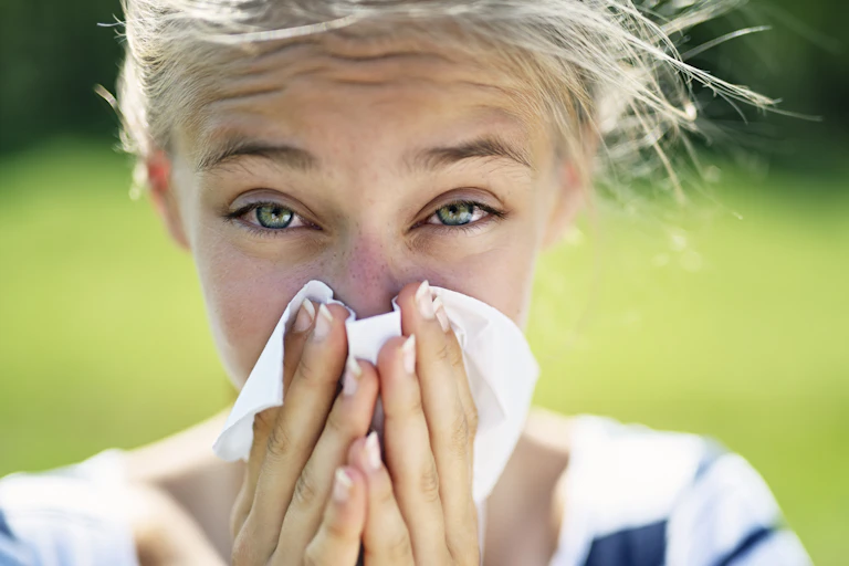 A young woman with allergy-reddened eyes sneezes into a tissue. The background is a lawn.