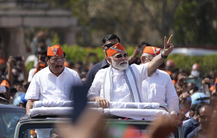 Indian prime minister Modi on the back of a car with two other men waving to a crowd of people.