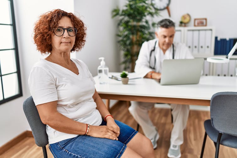 A woman facing the camera looking frustrated with a doctor at his desk in the background looking at his computer screen