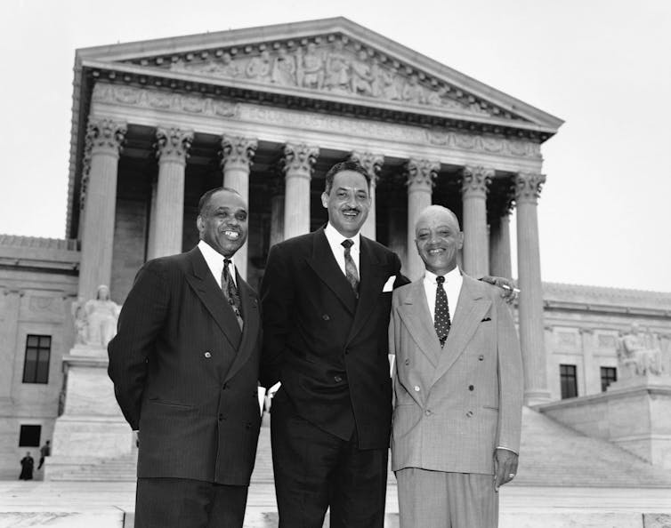 Three men in suits stand in front of the Supreme Court building.