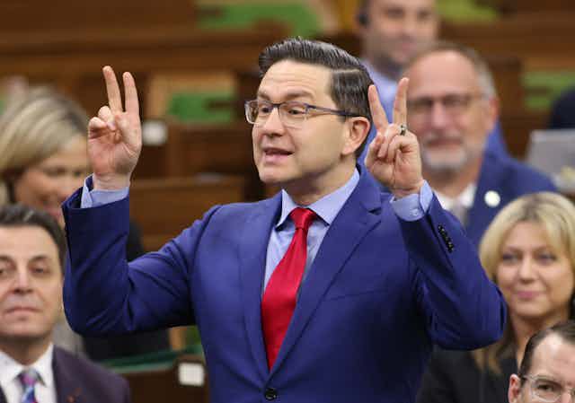 A man with dark air and glasses wearing a blue suit and red tie makes quote mark symbols with his hands.