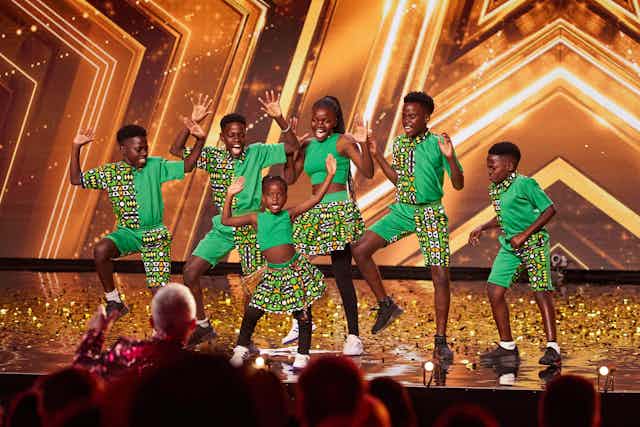 Six young dancers on a stage littered with gold streamers dressed in green outfits with African prints, hands in the air.