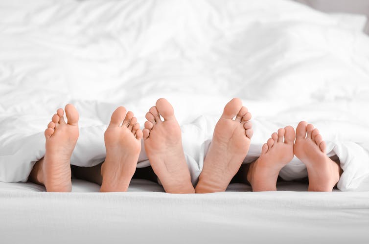 Three pairs of feet side by side, sticking out from under a white duvet