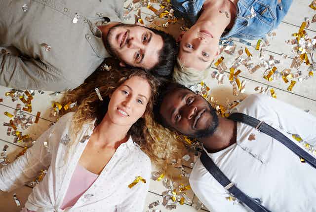 View from overhead of four young people lying with their heads together on a floor, with silver and gold confetti sprinkled over them
