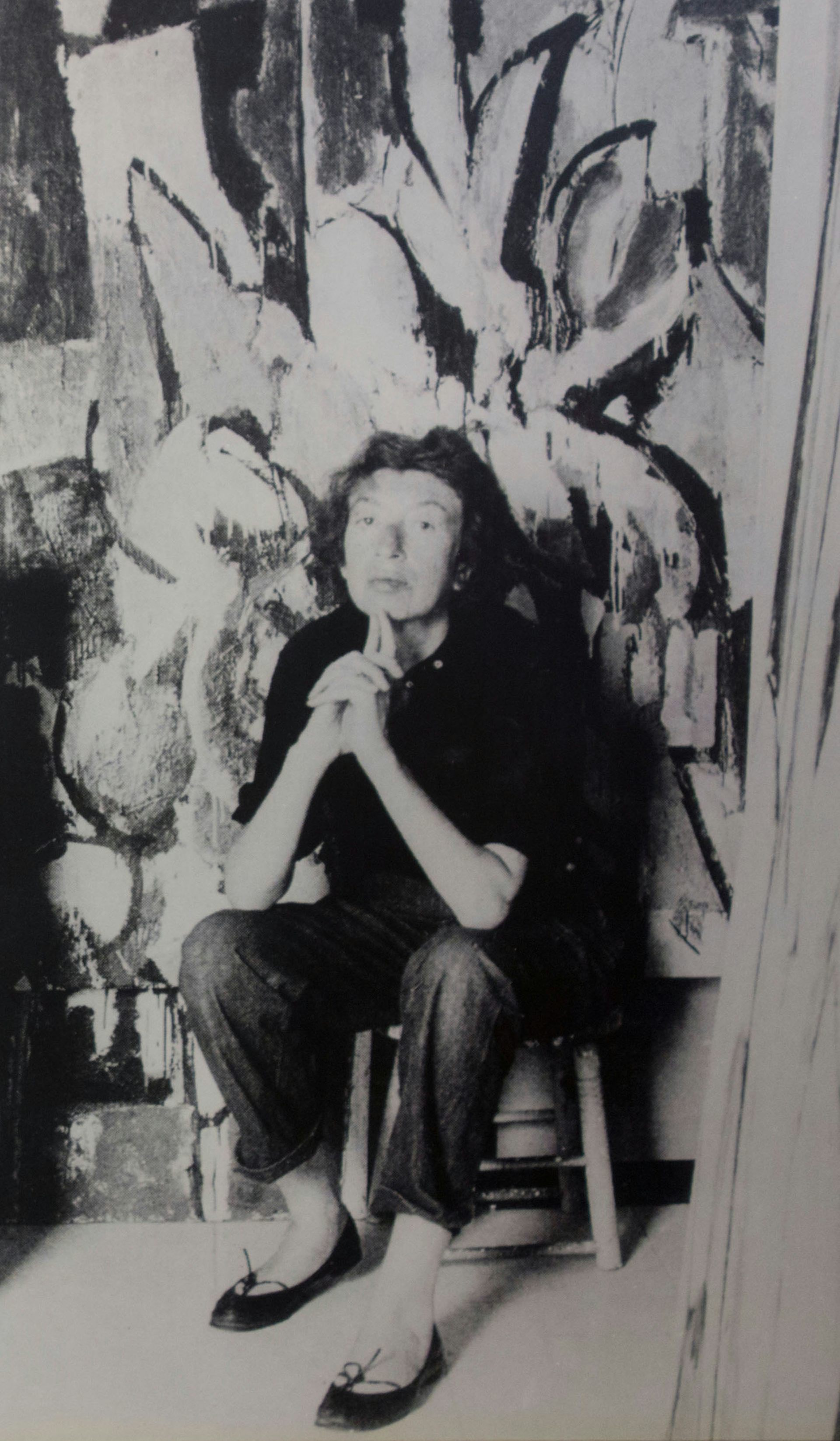 A woman dressed in black jumper and jeans sitting in front of an abstract expressionist painting.