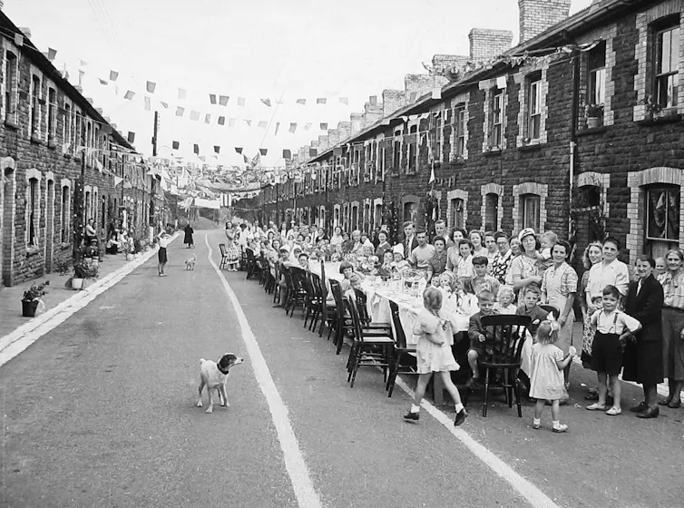 A black and white archival photograph of children at a long table in the street, with bunting overhead.